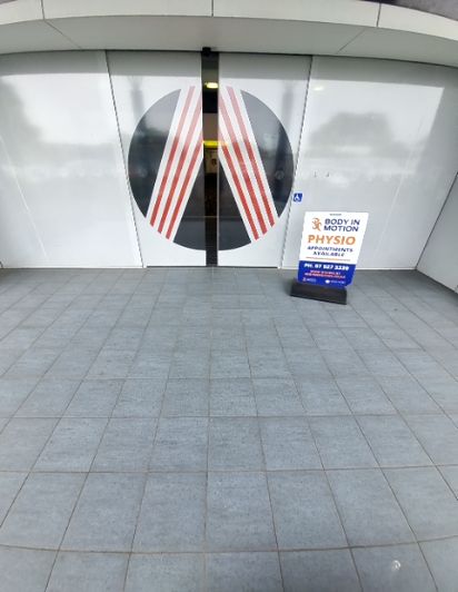 Level grey tiled pathway leading to automatic glass doors with a wheelchair symbol. A blue A-frame 'Physio' sign stands to the right of the entrance.