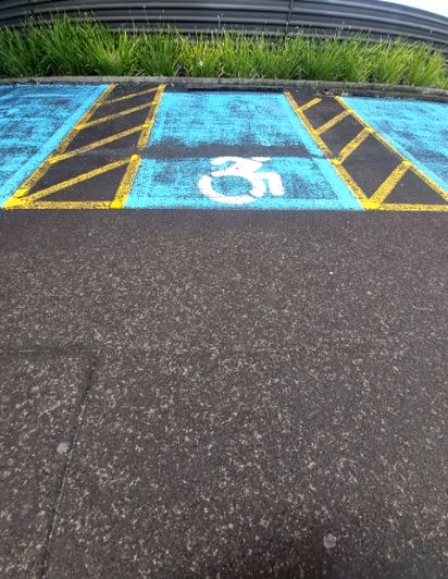 Blue accessible parking bay with white wheelchair symbol on asphalt. Adjacent access aisle marked with yellow lines and black diagonal stripes.
