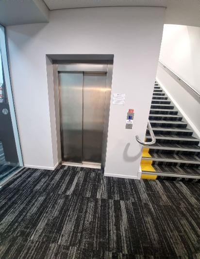 Stainless steel lift, black stairs with yellow visibility strips and silver handrail. Black and grey patterned carpeted floor.