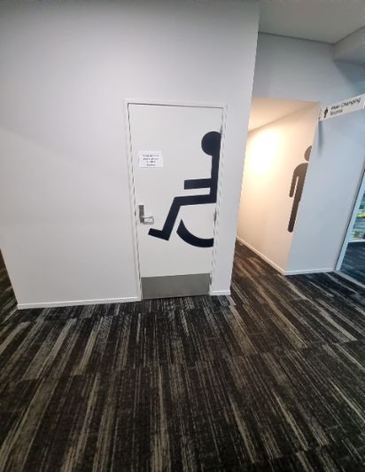 White door with large black wheelchair symbol for accessible toilet. Next to it, a hallway leads to 'Pee-Changing Rooms' with a standing person symbol. Dark striped carpeted floor.