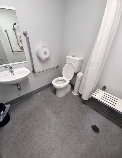 Accessible bathroom with a roll-in shower, foldable seat, and grab bars around the toilet and sink. Level, grey non-slip floor with a shower drain.