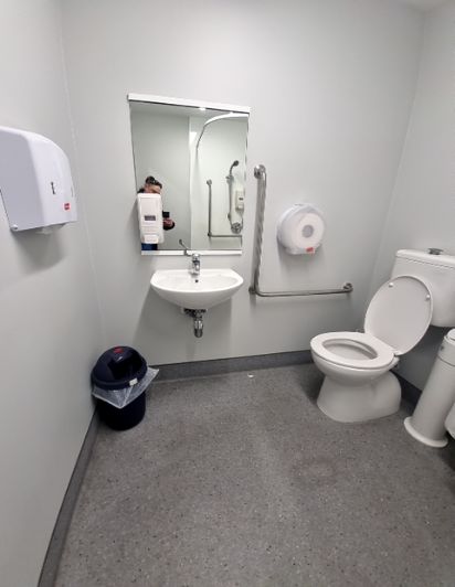 White toilet with silver grab bars. Wall-mounted sink. Shower area, also with grab bars, visible in mirror. Non-slip speckled grey floor. Hand dryer & toilet paper dispenser.