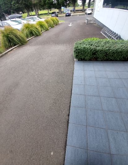 Level blue-grey tiled path next to a low hedge, bordering an asphalt driveway to a car park. Concrete steps and bicycle racks are visible near a white building.