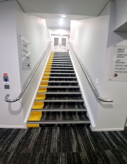 Carpeted staircase with yellow contrast nosings and handrails on both sides, leading to double doors labelled 'ATHLETES LOUNGE'. Emergency call point on left wall. Dark patterned carpet on ground.