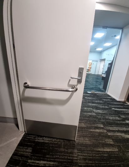 Partially open white door with silver lever handle, horizontal grab bar, and kick plate. Level carpeted flooring extends into a brightly lit hallway.