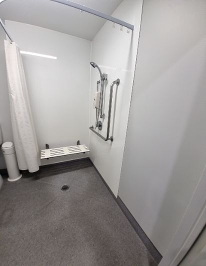 Roll-in shower with height-adjustable shower, grab bars, a foldable seat, and a level grey floor with drain.