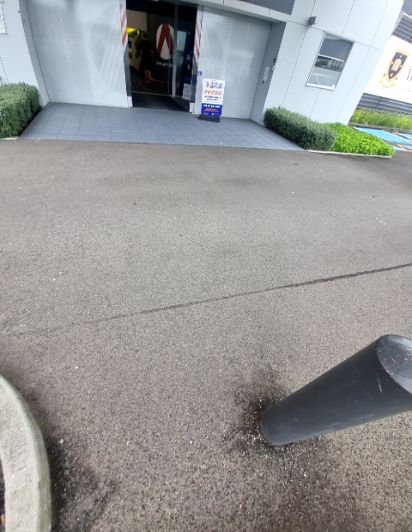 Level dark asphalt pathway leads to a wide building entrance with grey doors and a tiled forecourt. A blue sign and a grey bollard are visible.