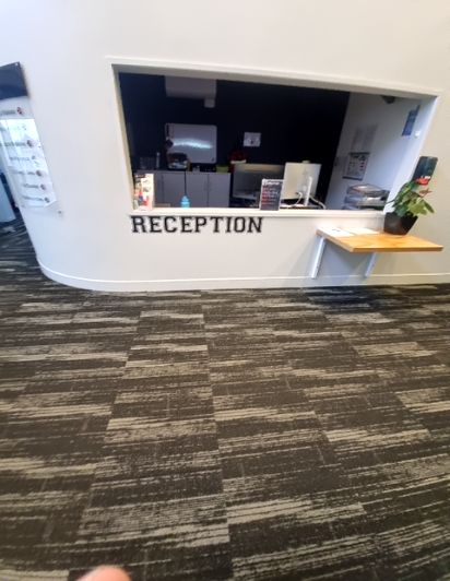 White reception desk with standard-height counter. A small wooden shelf is attached to the right. Level floor features dark grey patterned carpet tiles.