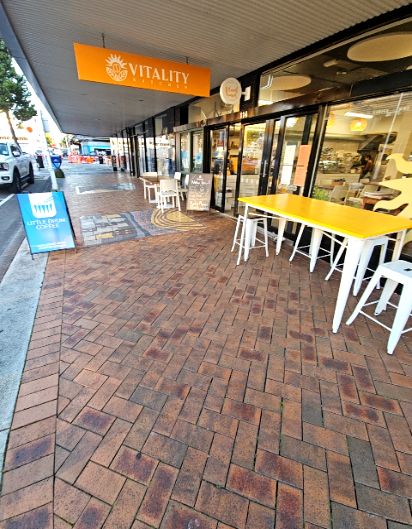 Level brick paved footpath leads to a wide glass shop entrance. Outdoor seating includes a long yellow table with white stools and white chairs. Clear overhead and blue A-frame signage.