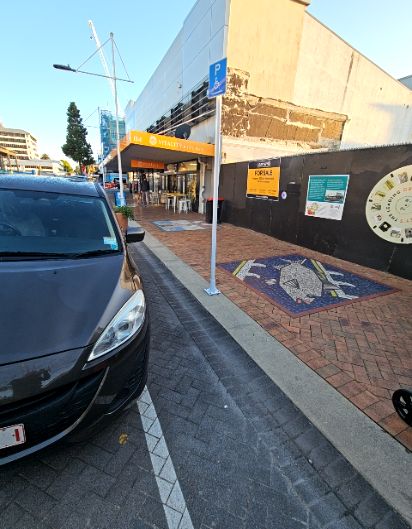 Blue accessible parking sign on a white pole beside a level red brick footpath. Path leads to a shop entrance with an orange awning. Black construction hoarding borders the right side of path.