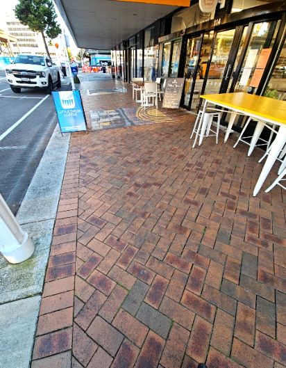 Level brick-paved footpath with herringbone pattern. Shop entrance has no visible step. Outdoor seating and an A-frame sign reduce walkway width. Concrete kerb borders the street.