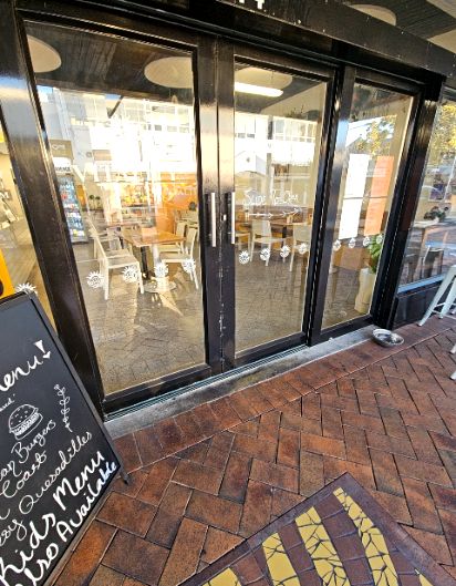 Level entry through glass sliding doors with black frames and silver handles. Red brick herringbone paving leads to a metal threshold. A menu chalkboard sits on the pavement to the left.