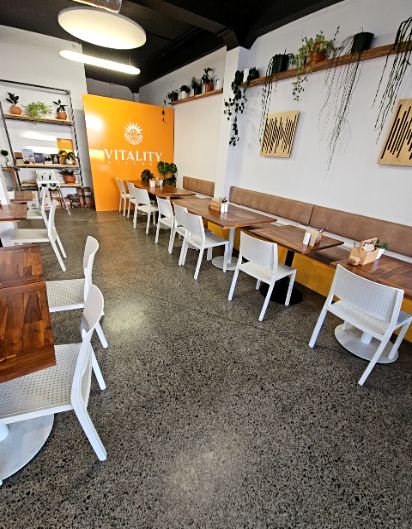 Level dark grey speckled concrete floor. Wooden tables with white armless chairs and a brown padded bench seat. Large orange wall with white signage and bright overhead lights.