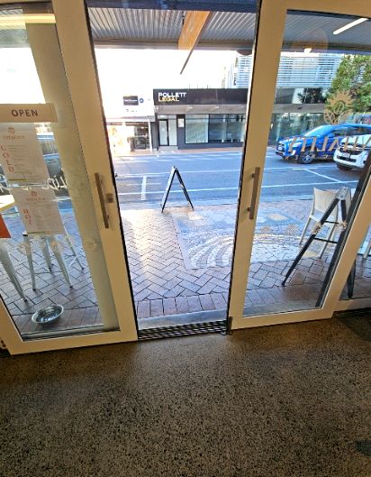 Level entrance via double glass doors with silver handles and a low metal threshold. Speckled grey floor meets a brick and mosaic footpath. A-frame signs sit on the wide path outside.