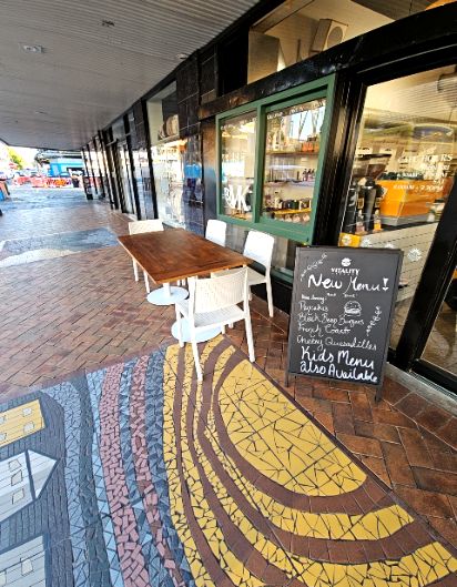 Level brick paved walkway with a decorative mosaic. Wooden table and white chairs sit outside a glass shopfront with a level entrance. A black A-frame sign stands on the wide, covered path.
