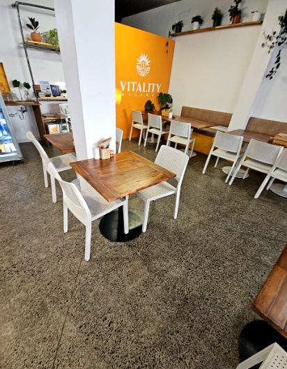 Level grey aggregate concrete floor with square timber tables and white armless chairs. An orange wall with signage and brown cushioned bench seating are visible. Bright indoor lighting.