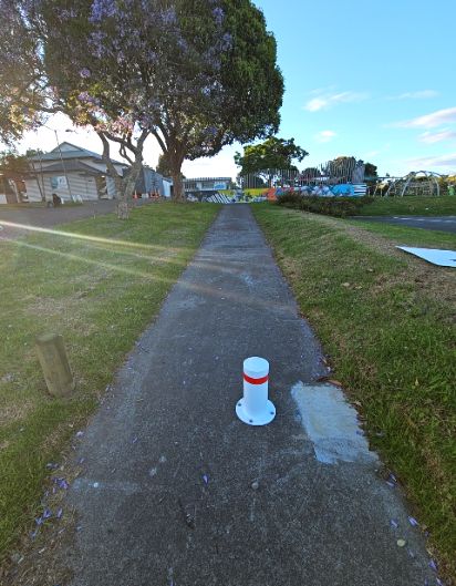 Sloping concrete pathway with a white and red bollard obstructing the centre. Grassy banks and a large tree line the path leading uphill towards a building and colourful mural.