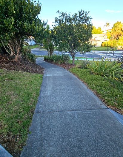 Wide grey concrete pathway with a gentle downhill gradient, bordered by grass and mulch garden beds, leading towards a road.