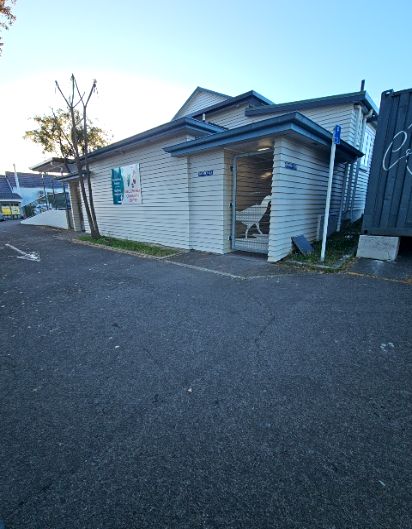 Level asphalt driveway leads to the gated entrance of a weatherboard community centre. A blue accessibility sign is on a pole nearby. A sign on the wall reads 'Community Centre'.