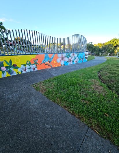 Wide level concrete pathway curves alongside a low wall with a colourful floral mural and wavy vertical metal fence. A grassy lawn borders the path on the right.