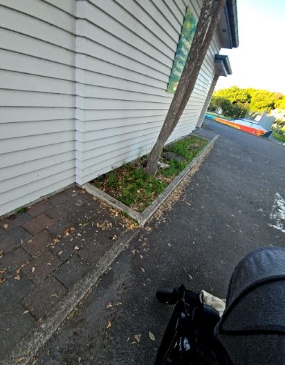 Brown brick path beside a white weatherboard wall. A concrete kerb and garden bed with a leaning tree narrow the walkway where it meets a grey asphalt driveway.