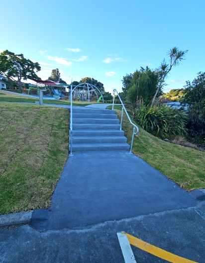 Six concrete steps with silver metal handrails on both sides lead from a dark grey asphalt pathway up to a light grey concrete path. Grass borders the steps under bright daylight.