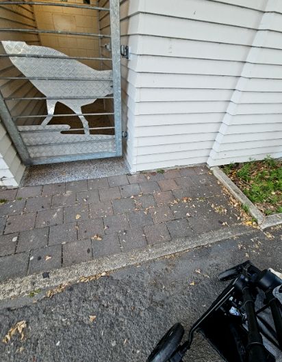 Metal gate with bird motif opens to a level speckled threshold. Approach via a grey brick path with a small concrete lip transition from asphalt. White weatherboard walls line the entrance.